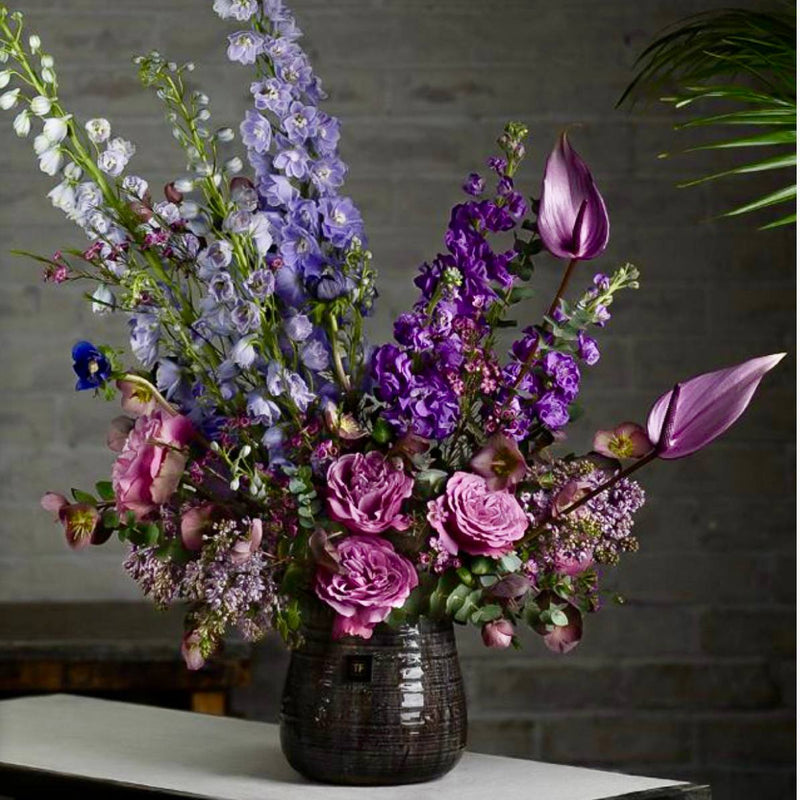 Purple and lavender Halloween arrangement with delphinium, anthurium, stock, and garden roses in a dark vessel for Los Angeles delivery.
