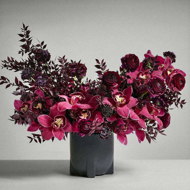 Modern wine burgundy Halloween centerpiece with magenta cymbidium orchids, deep red ranunculus, and scabiosa in a black vase for Los Angeles delivery.