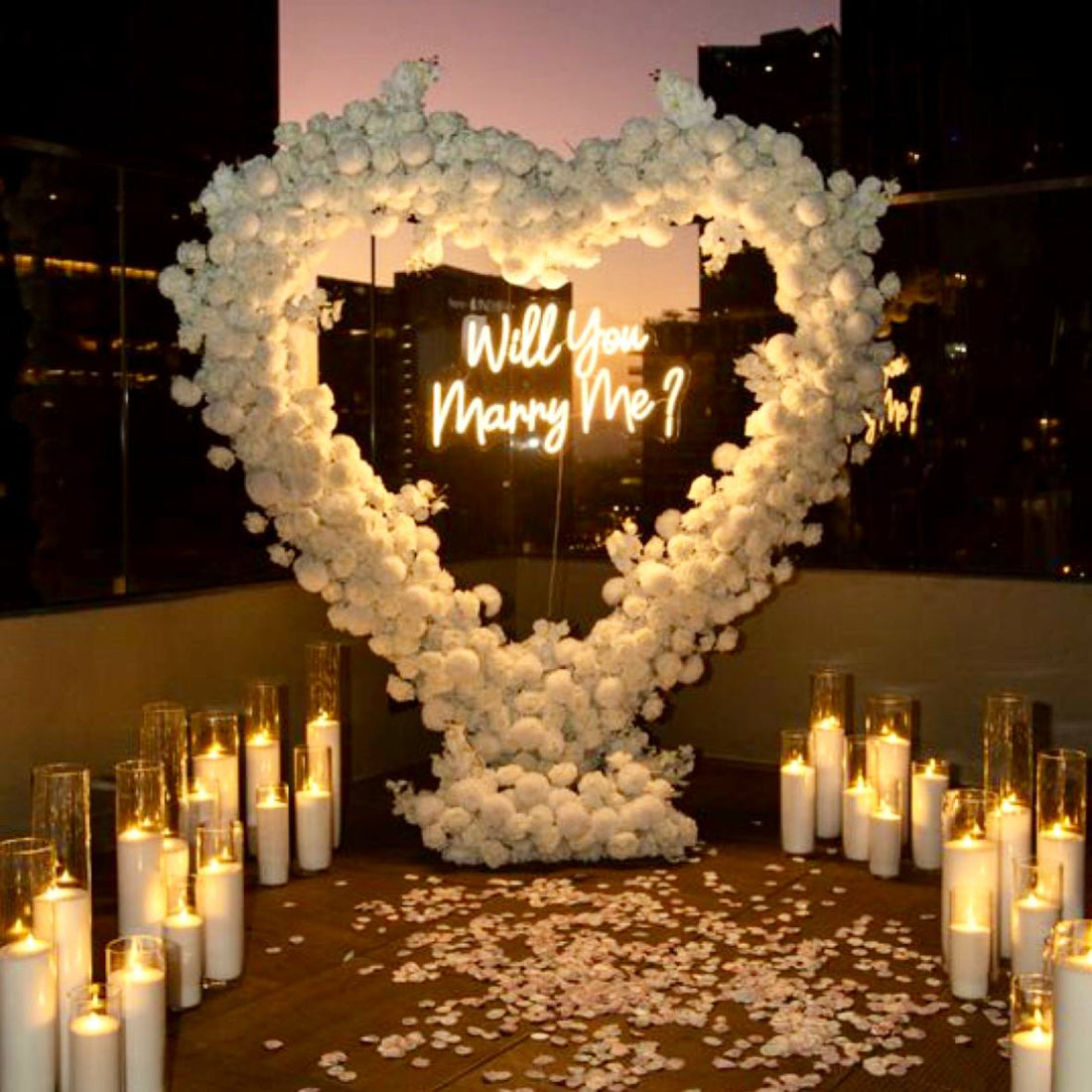 Heart-shaped ivory rose arch with Will You Marry Me neon sign, surrounded by pillar candles in glass cylinders and rose petals for Los Angeles proposal.