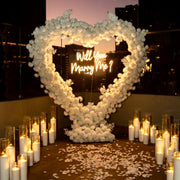 Heart-shaped ivory rose arch with Will You Marry Me neon sign, surrounded by pillar candles in glass cylinders and rose petals for Los Angeles proposal.