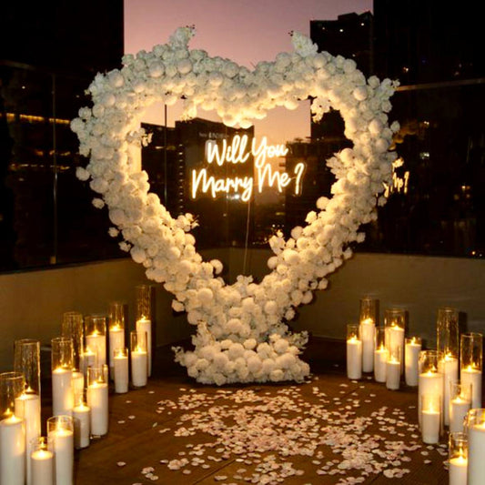 Heart-shaped ivory rose arch with Will You Marry Me neon sign, surrounded by pillar candles in glass cylinders and rose petals for Los Angeles proposal.