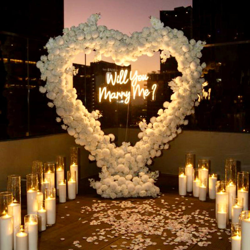 Heart-shaped ivory rose arch with Will You Marry Me neon sign, surrounded by pillar candles in glass cylinders and rose petals for Los Angeles proposal.
