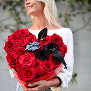 Dramatic Halloween bouquet featuring red roses, black blooms, feathers & raven decor in a heart-shaped keepsake box – ideal for luxury gifting