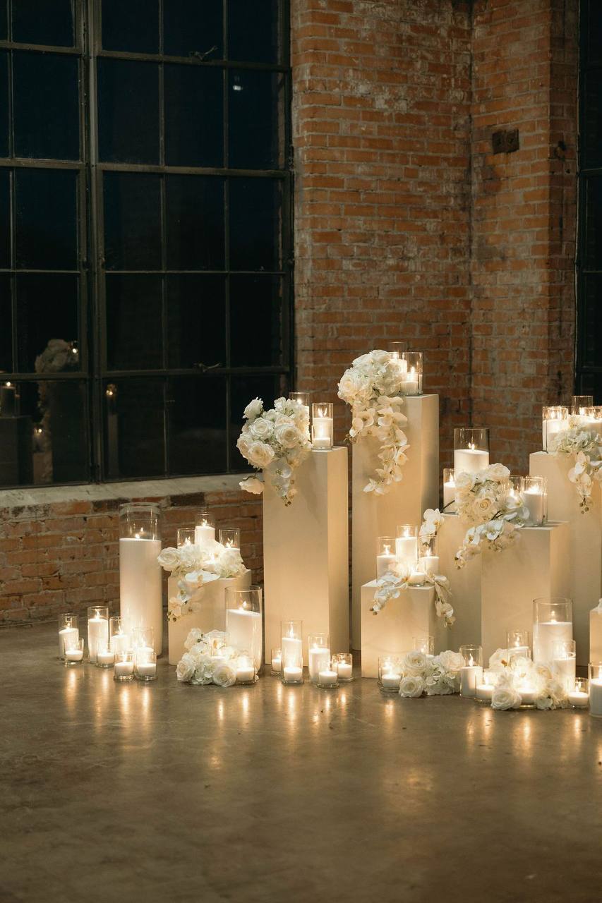 Industrial loft proposal setup with white rose and orchid pedestal florals, cascading orchids, and dozens of pillar candles against exposed brick wall in Los Angeles.