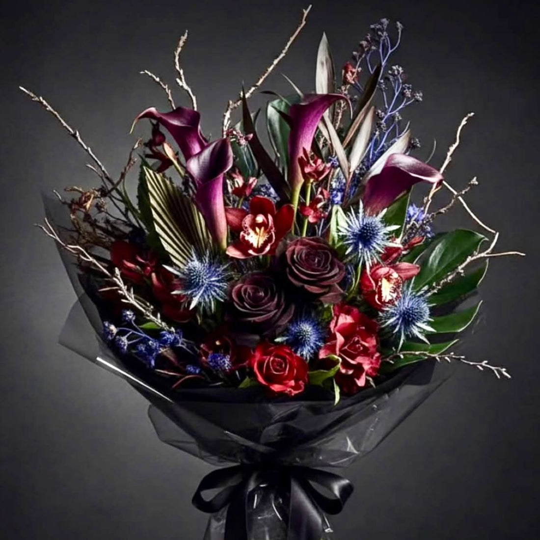 Dark Halloween hand-tied bouquet with black calla lilies, deep red cymbidium orchids, blue eryngium thistle, and twisting branches in black wrap.