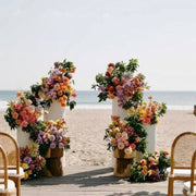 White pillar pedestals with pastel multi-color floral installations on a Los Angeles beach with bohemian rattan ceremony chairs.