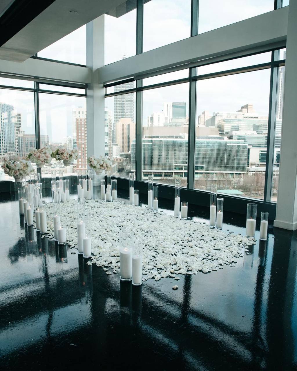 Penthouse proposal setup with twin floral pedestals, white rose petal aisle, and double row of glass cylinder candles with Los Angeles skyline view.