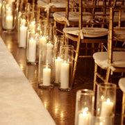 Pillar candles in tall glass hurricane cylinders lining a wedding aisle with gold chiavari chairs in Los Angeles.