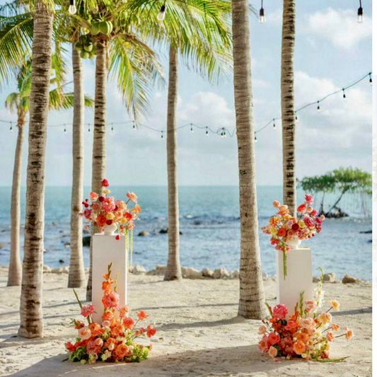 Tropical beach wedding with white pedestals featuring coral and peach floral arrangements under palm trees and string lights in Los Angeles.