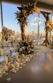 Rooftop proposal arch with wood frame, ivory roses, pampas grass, and Will You Marry Me neon sign overlooking the Los Angeles skyline at sunset.