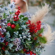 Festive floral arrangement showcasing silvery pinecones, blue thistle and lush greenery in a celebra